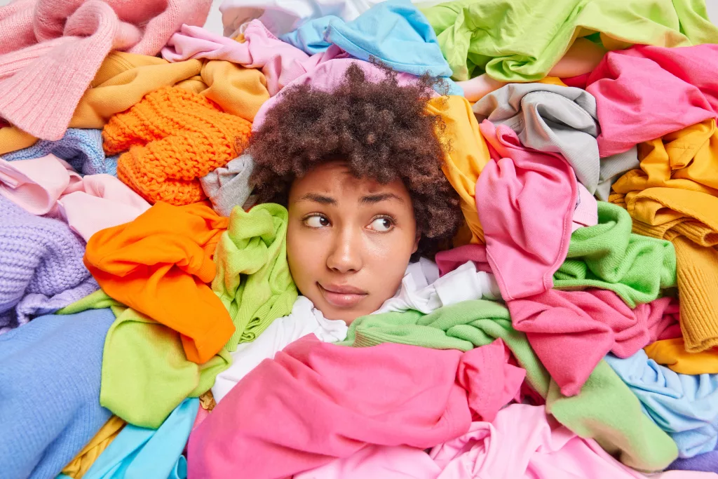 Curly haired woman collects clothes items in good condition to consignment shop or thrift store surrounded by huge piles of multicolored garments focused away has tired expression. Recycling textiles