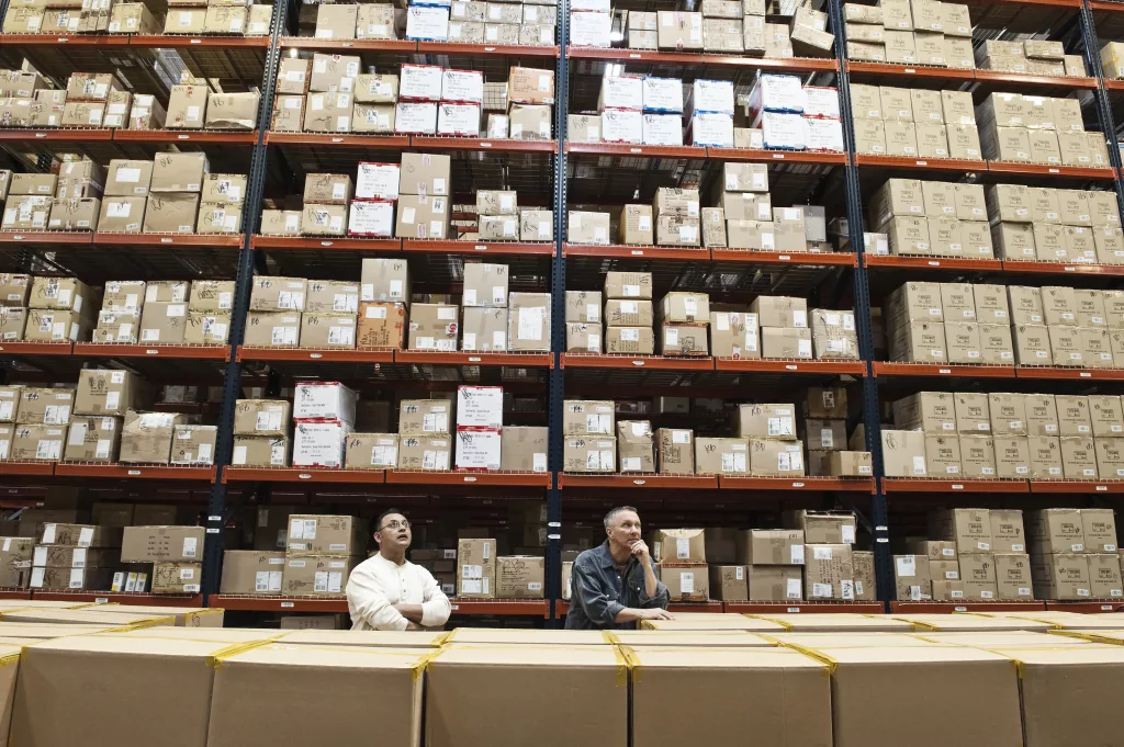 Two male warehouse workers checking inventory in a large distrubiton warehouse full of products stored in cardboard boxes on pallets on large racks.