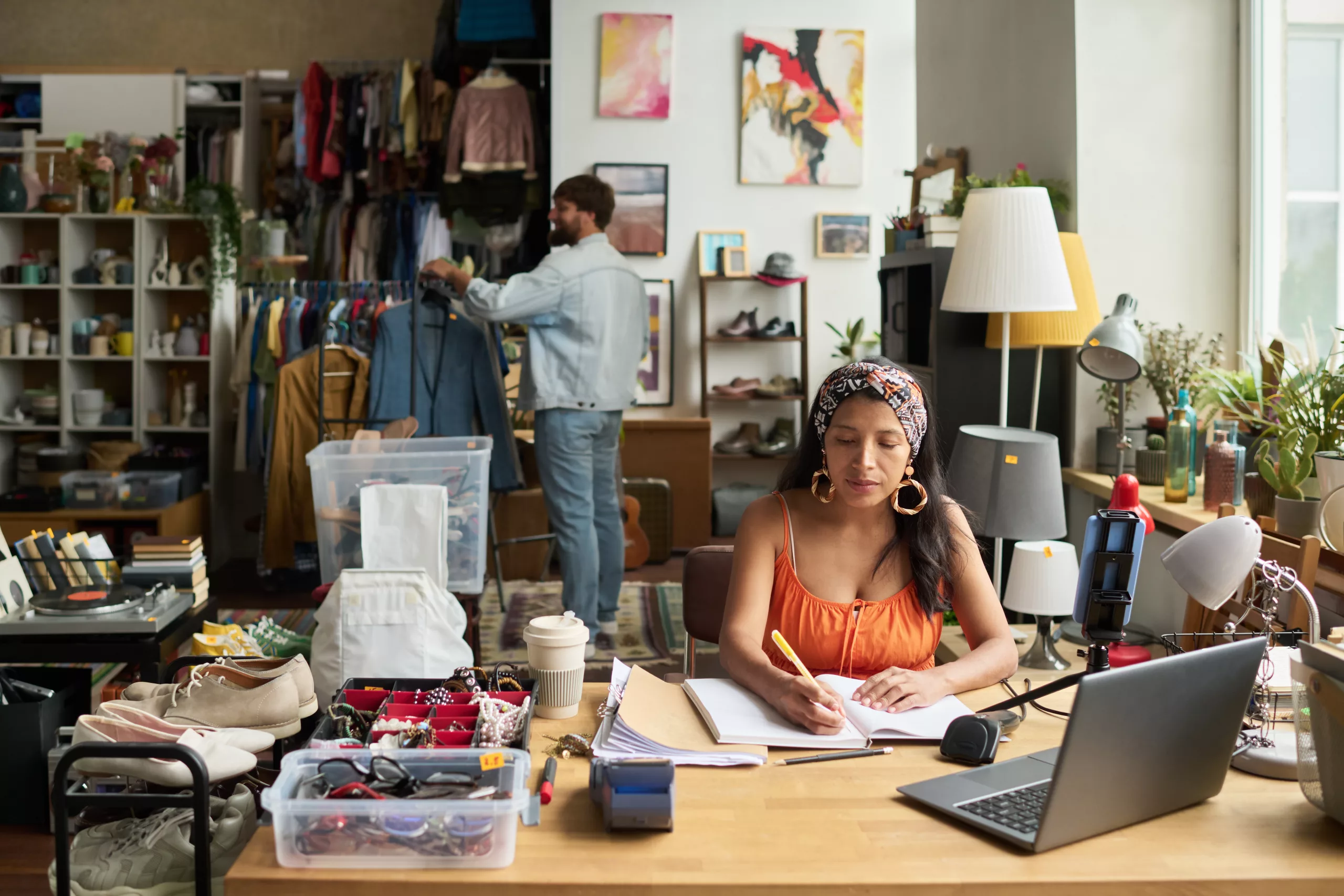 Young female owner of thrift shop sitting by workplace and making notes in copybook against male consumer choosing attire